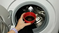 Červený hrnek s kávou před otevřenou pračkou, ruční vkládání tekutiny dovnitř.