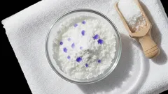 Prací prášek s modrými granulemi v misce, dřevěná lopatka na bílém ručníku.