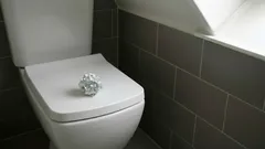 Moderní toaleta s ubrouskem na víku, šedé obklady, denní světlo z okna.